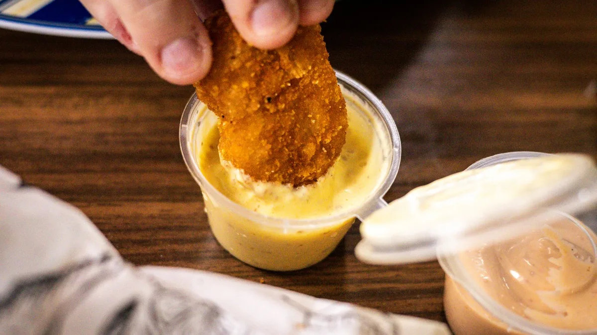 A crispy chicken bite being dipped into creamy sauce — Dave's Saucy Bites sauce on the side