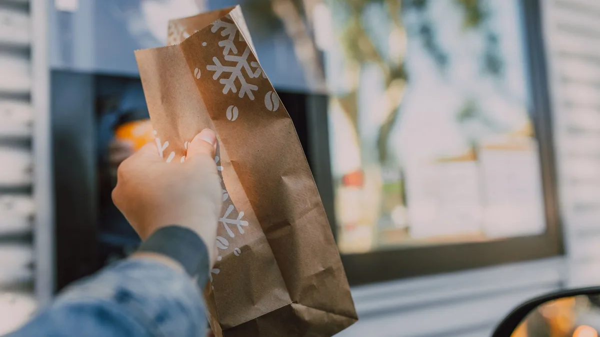 A hand reaching out with a paper takeout bag at a drive-thru window — Dave's Hot Chicken drive-thru
